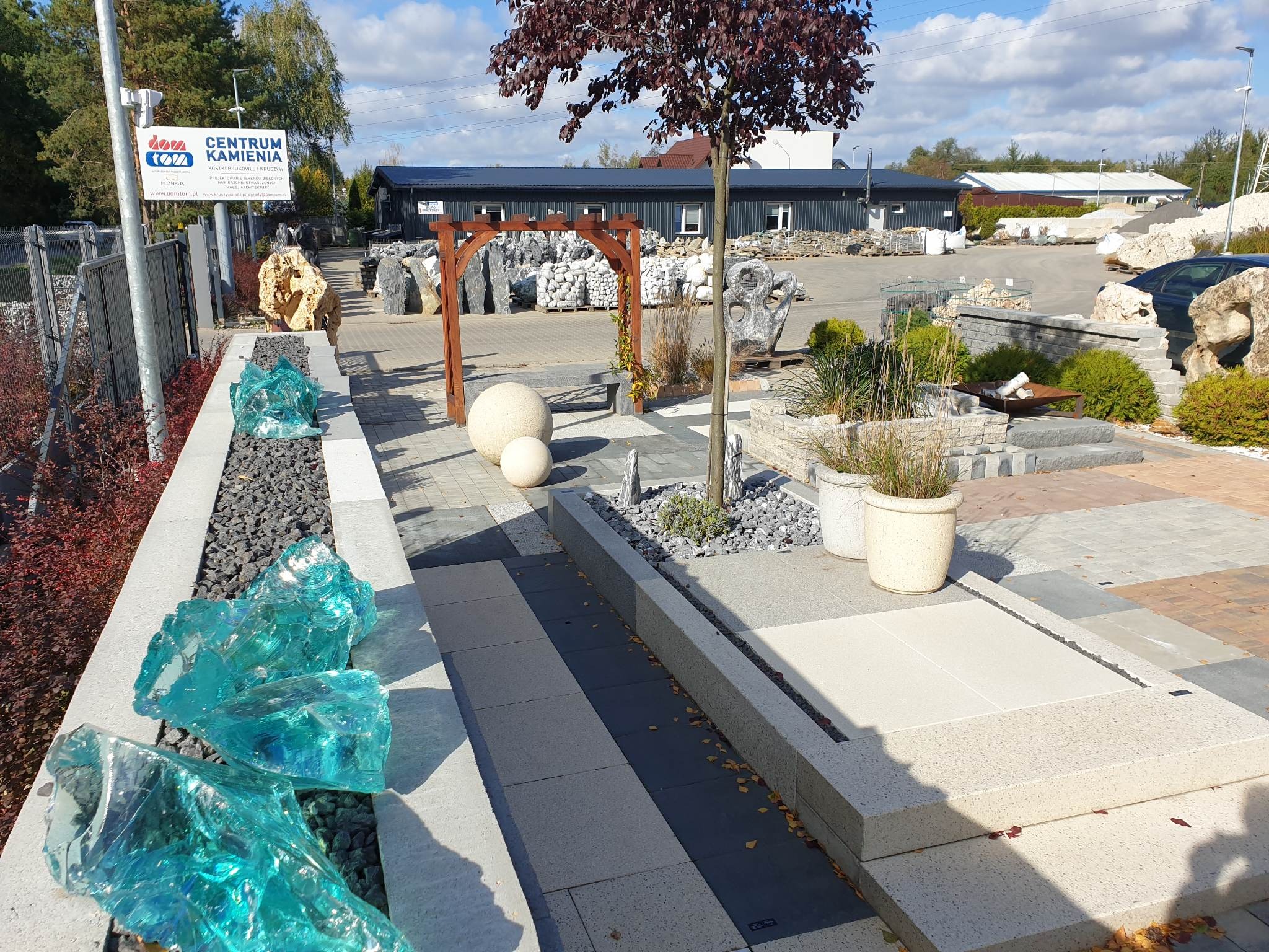 Ekspozycja różnorodnych materiałów nawierzchniowych: kostka brukowa, kamień ozdobny, żwir, drewniana pergola, betonowe kule i donice z roślinami. Widoczne elementy małej architektury w przestrzeni...