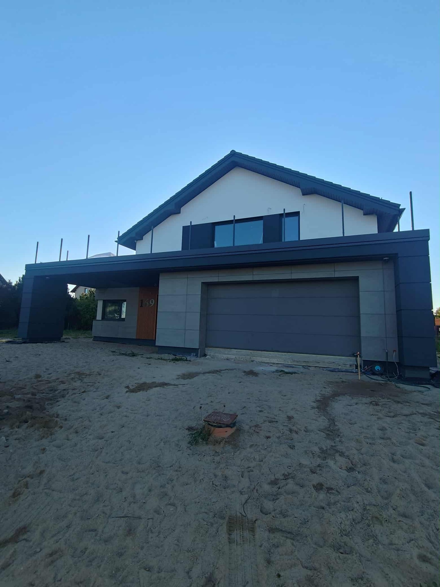 Nowoczesny dom jednorodzinny z szarą elewacją, widok z zewnątrz. Elementy wykończeniowe w kolorze antracytowym. Teren wokół domu w trakcie prac wykończeniowych.