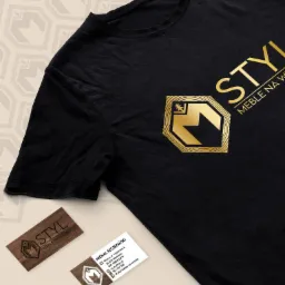 Czarny t-shirt z nadrukowanym złotym logo i nazwą firmy meblarskiej 'STYL Meble na Wymiar', obok dwie wizytówki z tym samym logo, na beżowym tle z powtarzającym się wzorem logo.