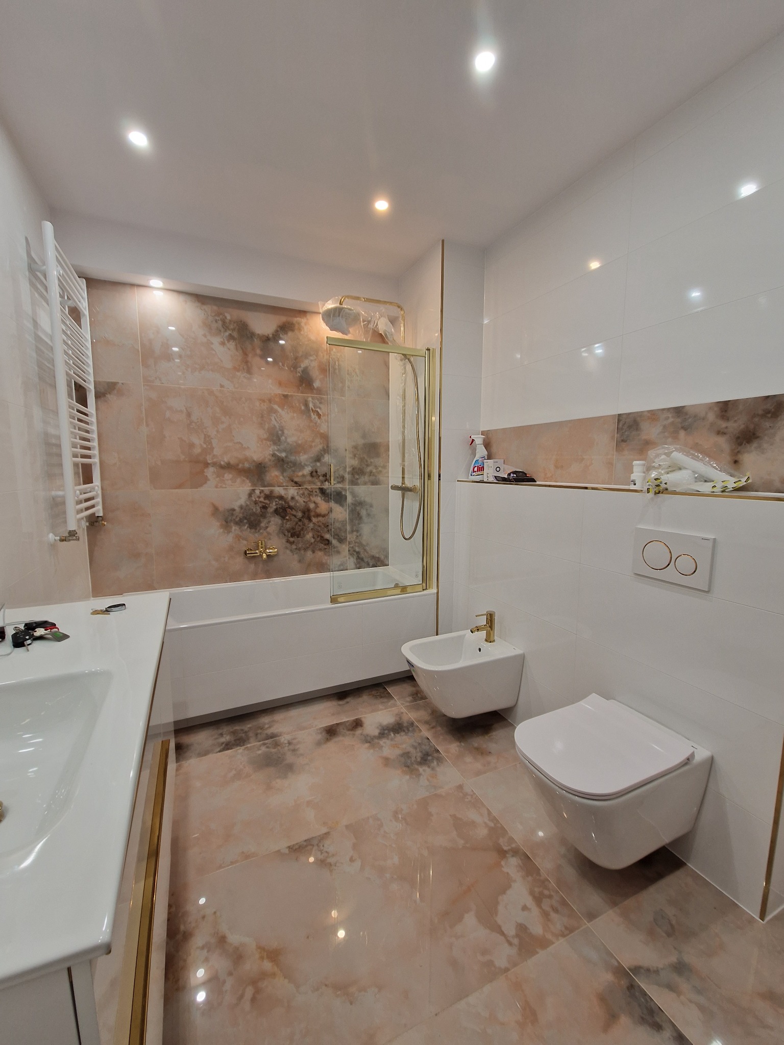 Nowoczesna łazienka po remoncie z marmurowymi płytkami, białą armaturą z złotymi akcentami i kabiną prysznicową ze złotymi ramami.