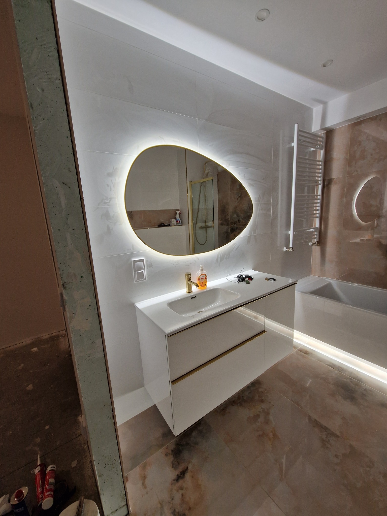 Nowoczesna łazienka w trakcie wykańczania, z podświetlanym lustrem o nieregularnym kształcie, białą szafką pod umywalką z złotymi akcentami, płytkami imitującymi marmur na podłodze i ścianach...