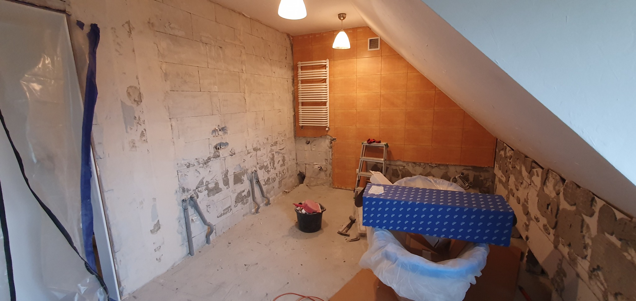 Łazienka w trakcie remontu: częściowo usunięte płytki, odsłonięte rury, zabezpieczona folią, widoczny grzejnik drabinkowy, drabina i narzędzia.