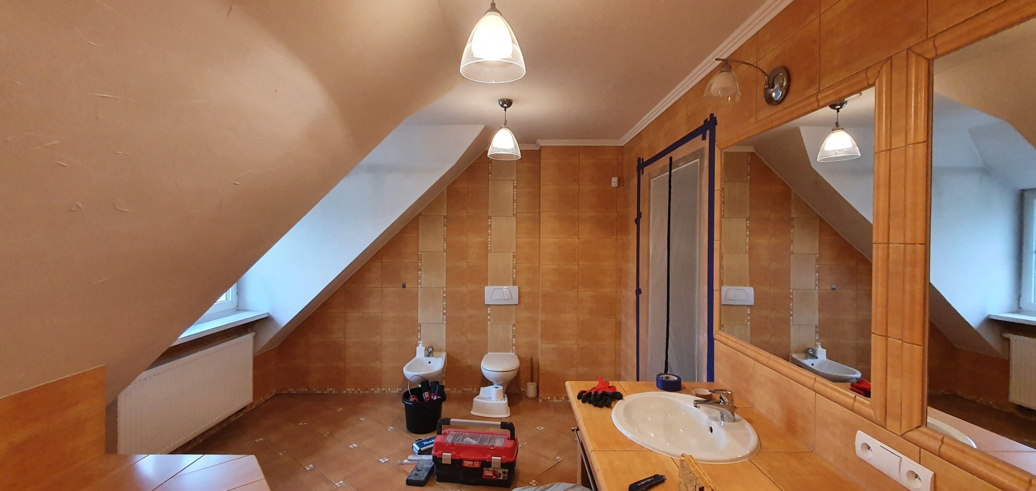Remontowana łazienka na poddaszu z brązowymi płytkami, widoczna umywalka, bidet, toaleta, grzejnik i narzędzia w kuwecie, drzwi zabezpieczone taśmą malarską.