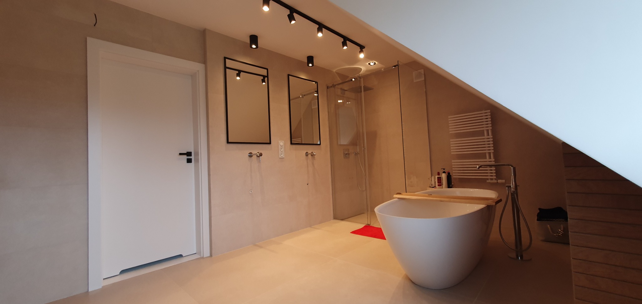 Nowoczesna łazienka na poddaszu ze skośnym sufitem, wolnostojąca biała wanna z drewnianą deską, szklana kabina prysznicowa, dwa lustra w czarnych ramach, grzejnik drabinkowy i oświetlenie punktowe.