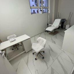 Mmremont Kamil Iskra - Jasne wnętrze gabinetu kosmetycznego z białymi meblami, marmurową posadzką i regulowanym fotelem do zabiegów. Na biurku leżą dokumenty.
