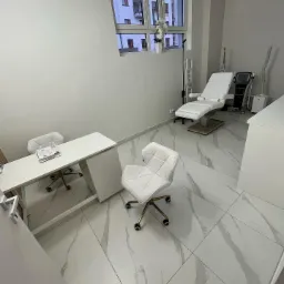 Jasne wnętrze gabinetu kosmetycznego z białymi meblami, marmurową posadzką i regulowanym fotelem do zabiegów. Na biurku leżą dokumenty.