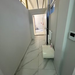 Mmremont Kamil Iskra - Korytarz wykończony w szarościach i bieli, z marmurową podłogą i widokiem na biuro w oddali, oświetlony naturalnym światłem wpadającym przez okno.