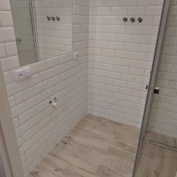 Mmremont Kamil Iskra - Wykończona łazienka z białymi, prostokątnymi płytkami na ścianach, imitującymi cegłę, oraz podłogą z płytek drewnopodobnych. Widoczna kabina prysznicowa z odpływem liniowym, lustro i chromowane...