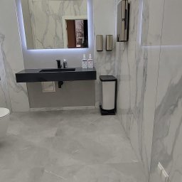 Mmremont Kamil Iskra - Nowoczesna łazienka z marmurowymi płytkami, czarną umywalką nablatową i podświetlanym lustrem, uzupełniona o srebrne dozowniki i biały kosz na śmieci.