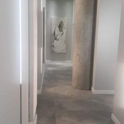Mmremont Kamil Iskra - Długi korytarz w minimalistycznym stylu z szarą podłogą imitującą kamień, białymi ścianami z wbudowanym oświetleniem LED, betonowym filarem z zielonym znakiem ewakuacyjnym i obrazem na końcu...