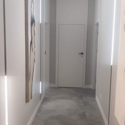Mmremont Kamil Iskra - Nowoczesny korytarz z szarą, marmurkową podłogą, białymi ścianami z wbudowanymi pionowymi lampami LED i obrazem w beżowej ramie, czarny sufit rastrowy.