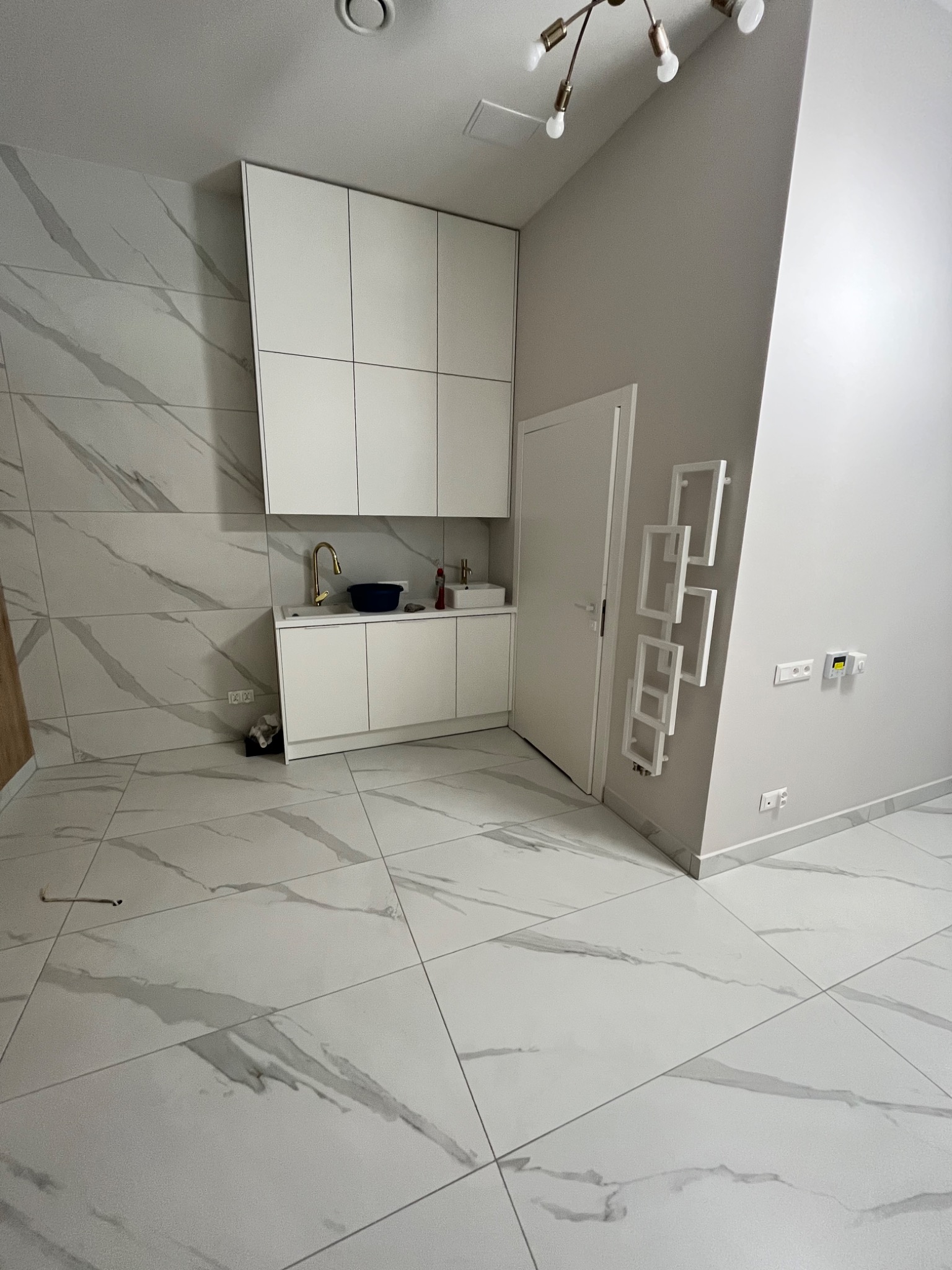 Nowoczesna, minimalistyczna łazienka z białymi płytkami imitującymi marmur na podłodze i ścianach, białe szafki pod umywalką z baterią w kolorze złota, oraz biała wisząca szafka. Na ścianie...