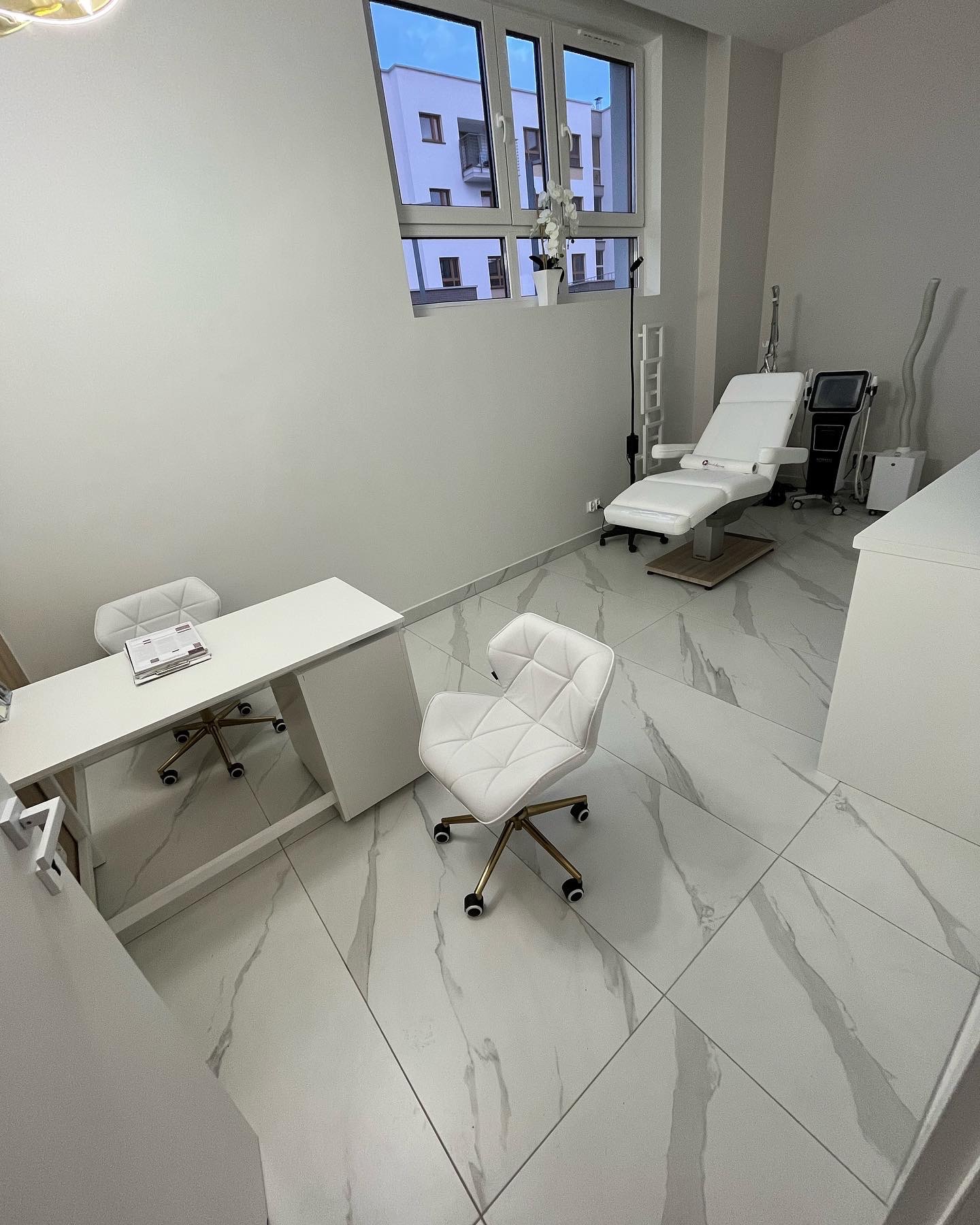 Jasne wnętrze gabinetu kosmetycznego z białymi meblami, marmurową posadzką i regulowanym fotelem do zabiegów. Na biurku leżą dokumenty.