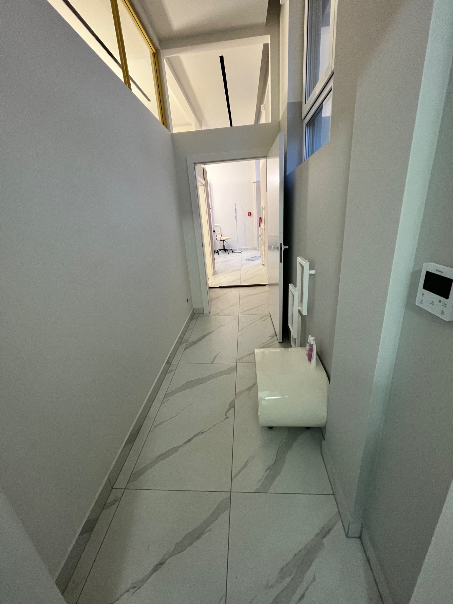 Korytarz wykończony w szarościach i bieli, z marmurową podłogą i widokiem na biuro w oddali, oświetlony naturalnym światłem wpadającym przez okno.
