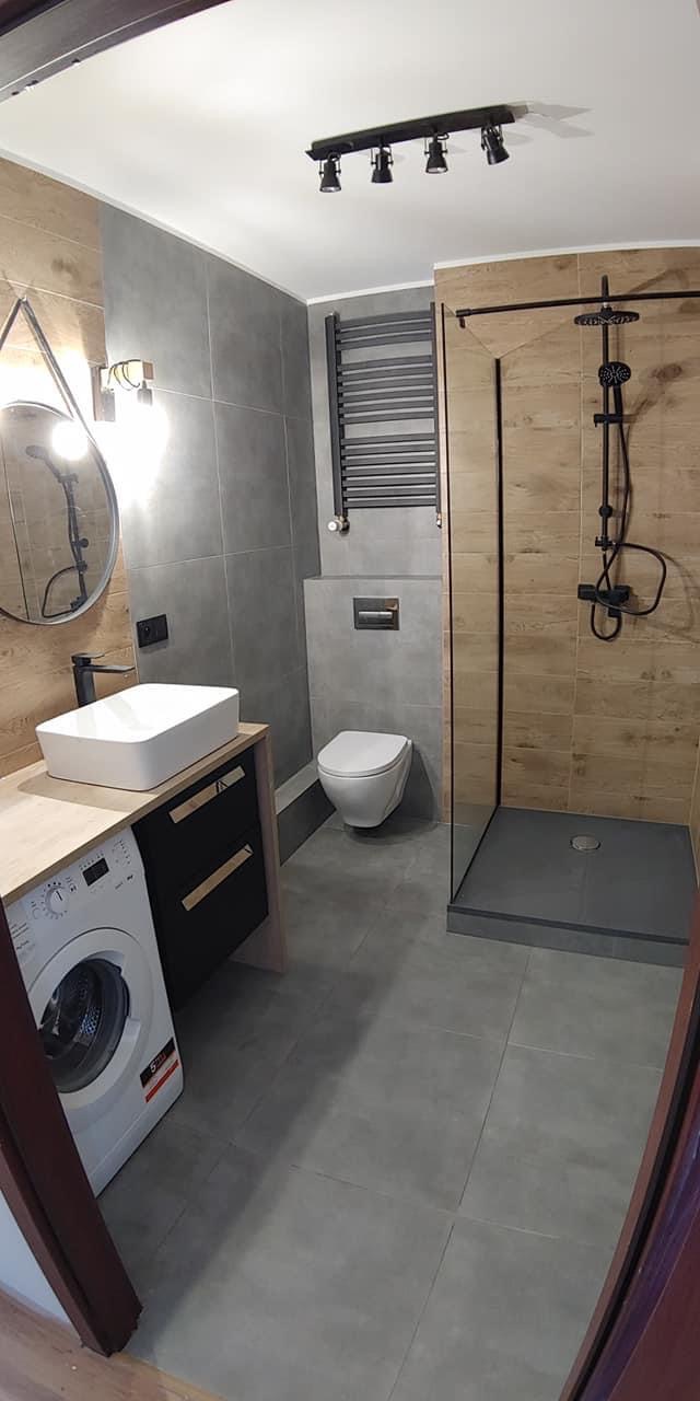 Nowoczesna łazienka z drewnopodobnymi i szarymi płytkami, kabiną prysznicową typu walk-in z czarną armaturą, umywalką nablatową, pralką i grzejnikiem drabinkowym w kolorze antracytowym.