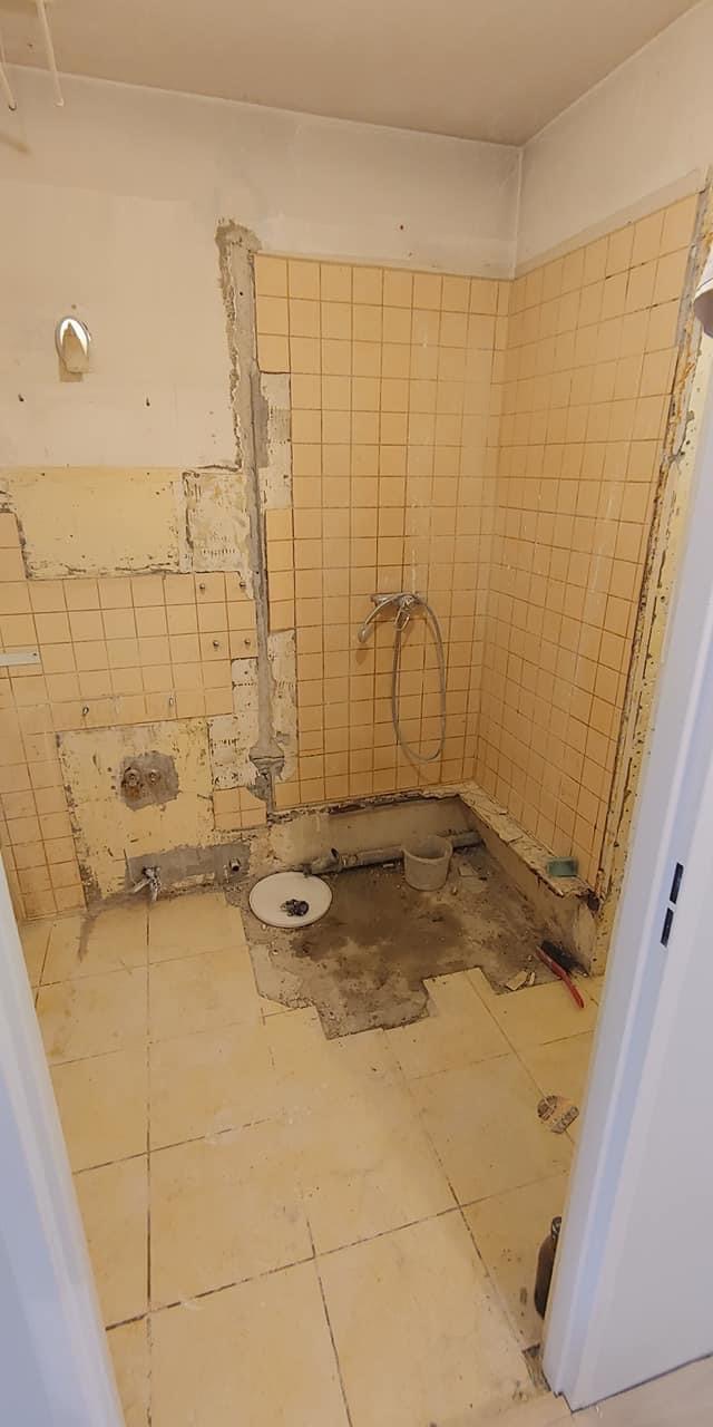 Demontaż płytek w łazience z odsłoniętymi rurami, widoczne pozostałości kleju i stara instalacja prysznicowa.