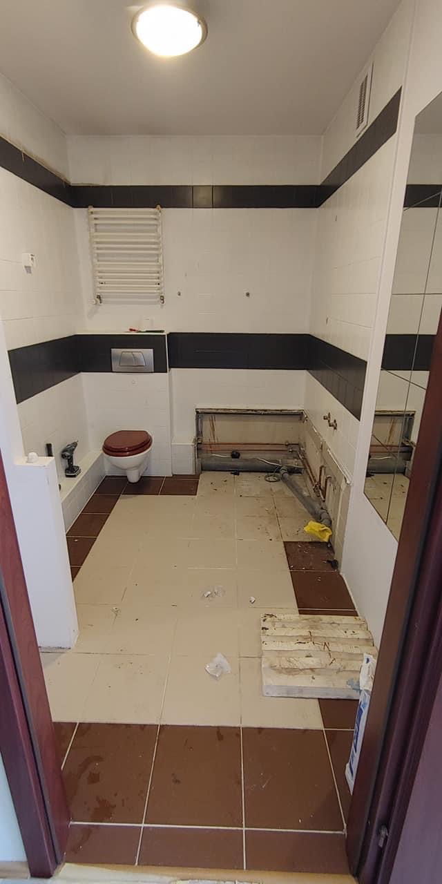 Łazienka w trakcie demontażu, widoczne fragmenty starej instalacji, częściowo usunięte płytki, toaleta z brązową deską, grzejnik drabinkowy na ścianie.