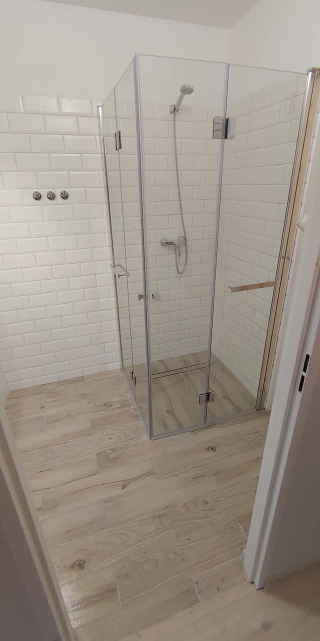 Kabina prysznicowa z transparentnego szkła, białe płytki typu metro na ścianach, drewnopodobna podłoga, trzy okrągłe wieszaki na ręczniki po lewej stronie.