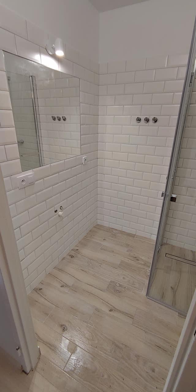 Wykończona łazienka z białymi, prostokątnymi płytkami na ścianach, imitującymi cegłę, oraz podłogą z płytek drewnopodobnych. Widoczna kabina prysznicowa z odpływem liniowym, lustro i chromowane...