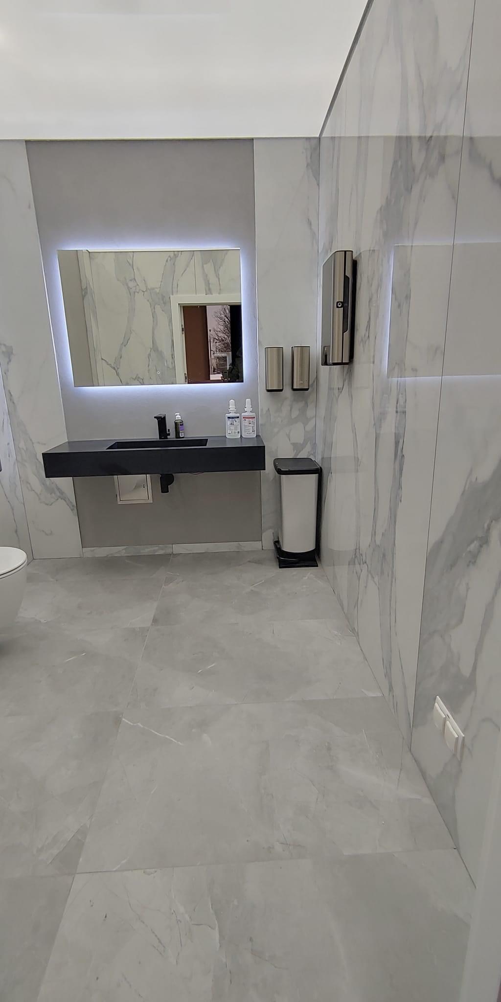 Nowoczesna łazienka z marmurowymi płytkami, czarną umywalką nablatową i podświetlanym lustrem, uzupełniona o srebrne dozowniki i biały kosz na śmieci.