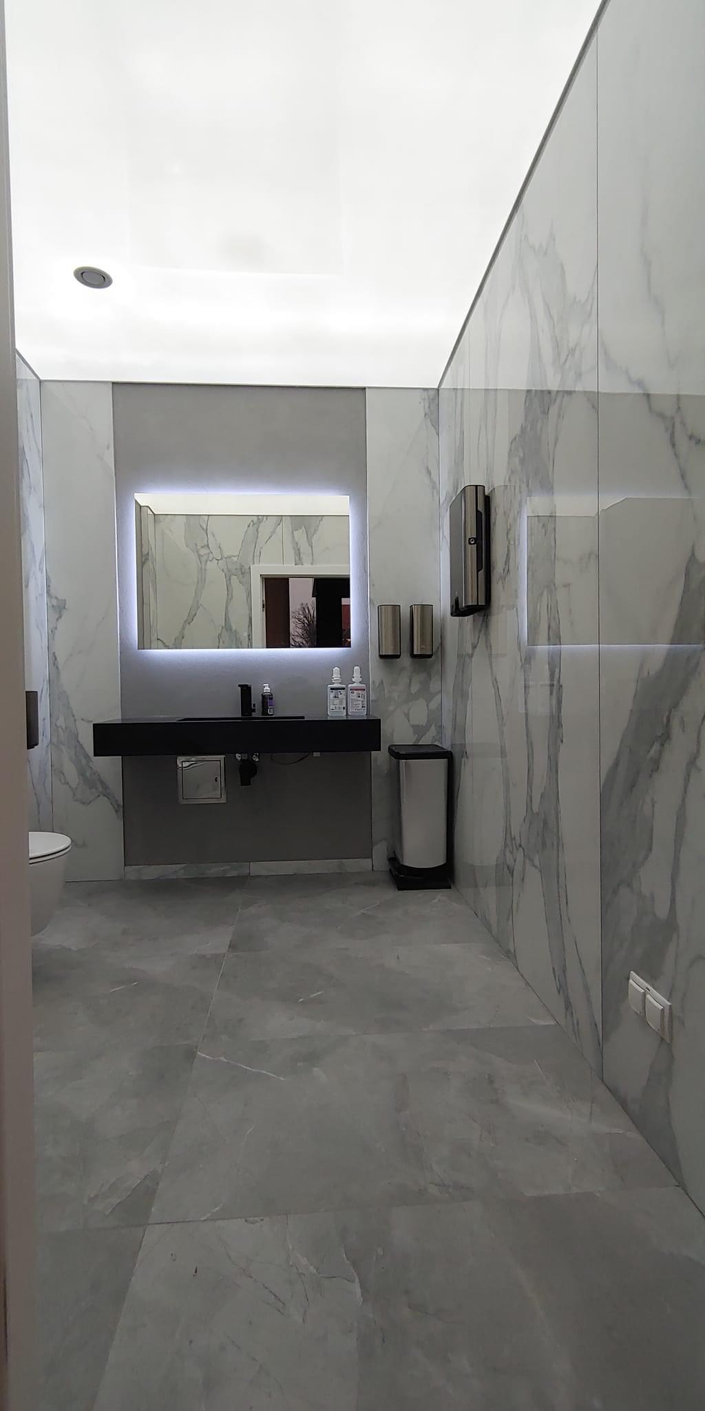 Nowoczesna łazienka w szarościach z podświetlanym lustrem, marmurowymi płytkami na ścianach i sufitem napinanym z oświetleniem punktowym.