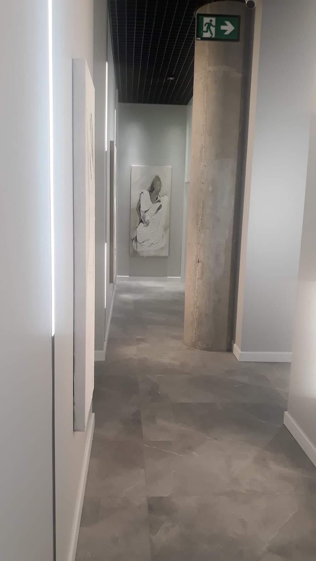 Długi korytarz w minimalistycznym stylu z szarą podłogą imitującą kamień, białymi ścianami z wbudowanym oświetleniem LED, betonowym filarem z zielonym znakiem ewakuacyjnym i obrazem na końcu...