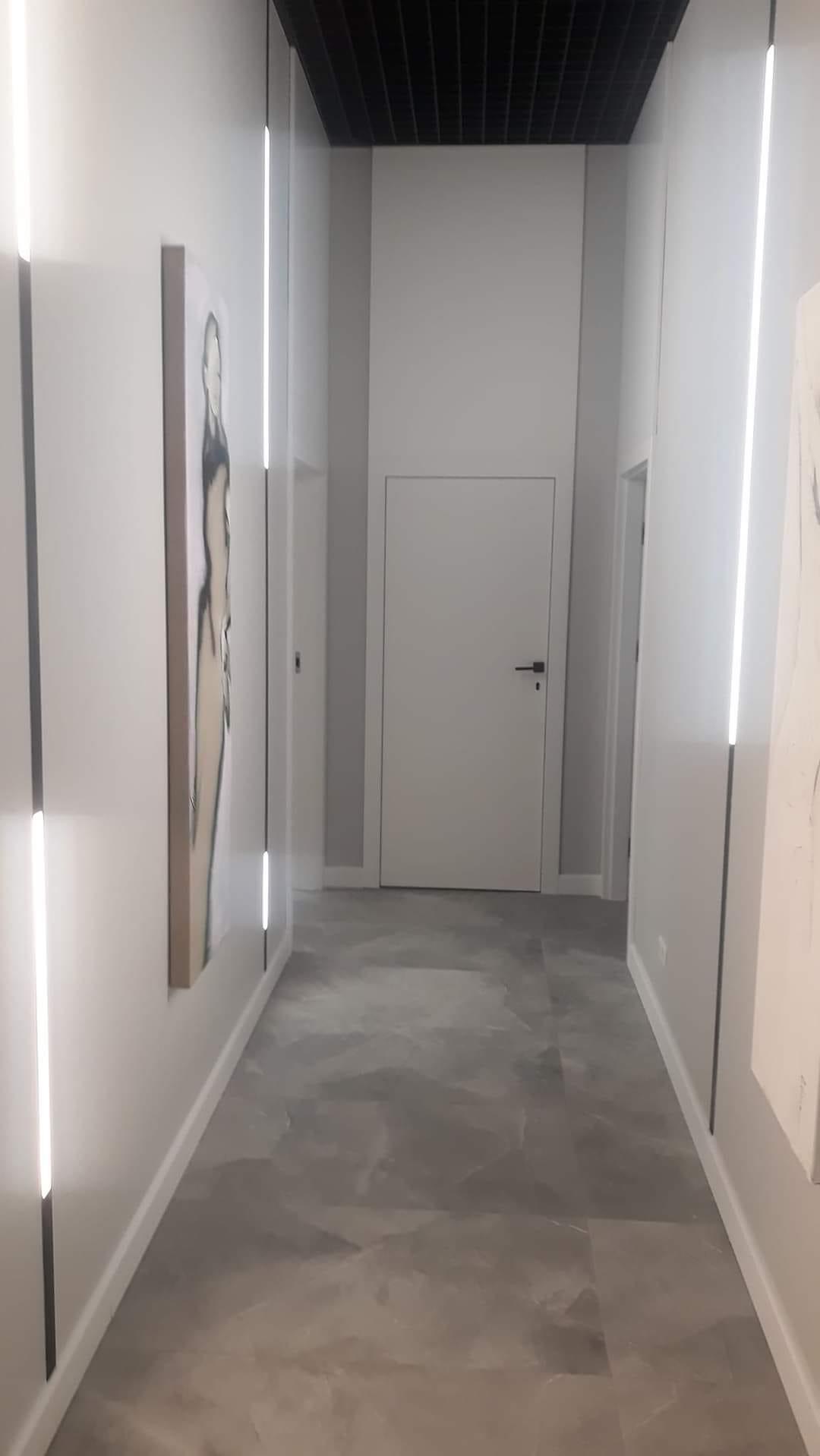 Nowoczesny korytarz z szarą, marmurkową podłogą, białymi ścianami z wbudowanymi pionowymi lampami LED i obrazem w beżowej ramie, czarny sufit rastrowy.