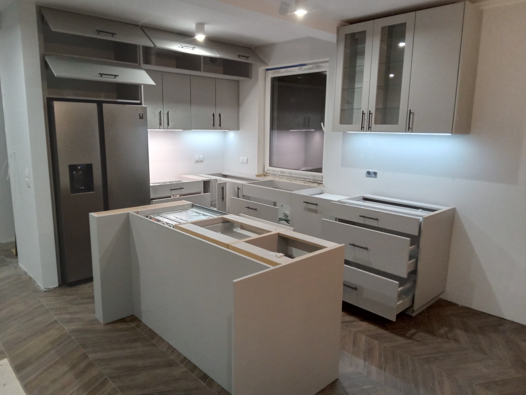 Montaż szarej kuchni na wymiar z wyspą kuchenną, widoczne otwarte szafki wiszące i szuflady w trakcie instalacji, na podłodze ułożona parkiet w jodełkę.