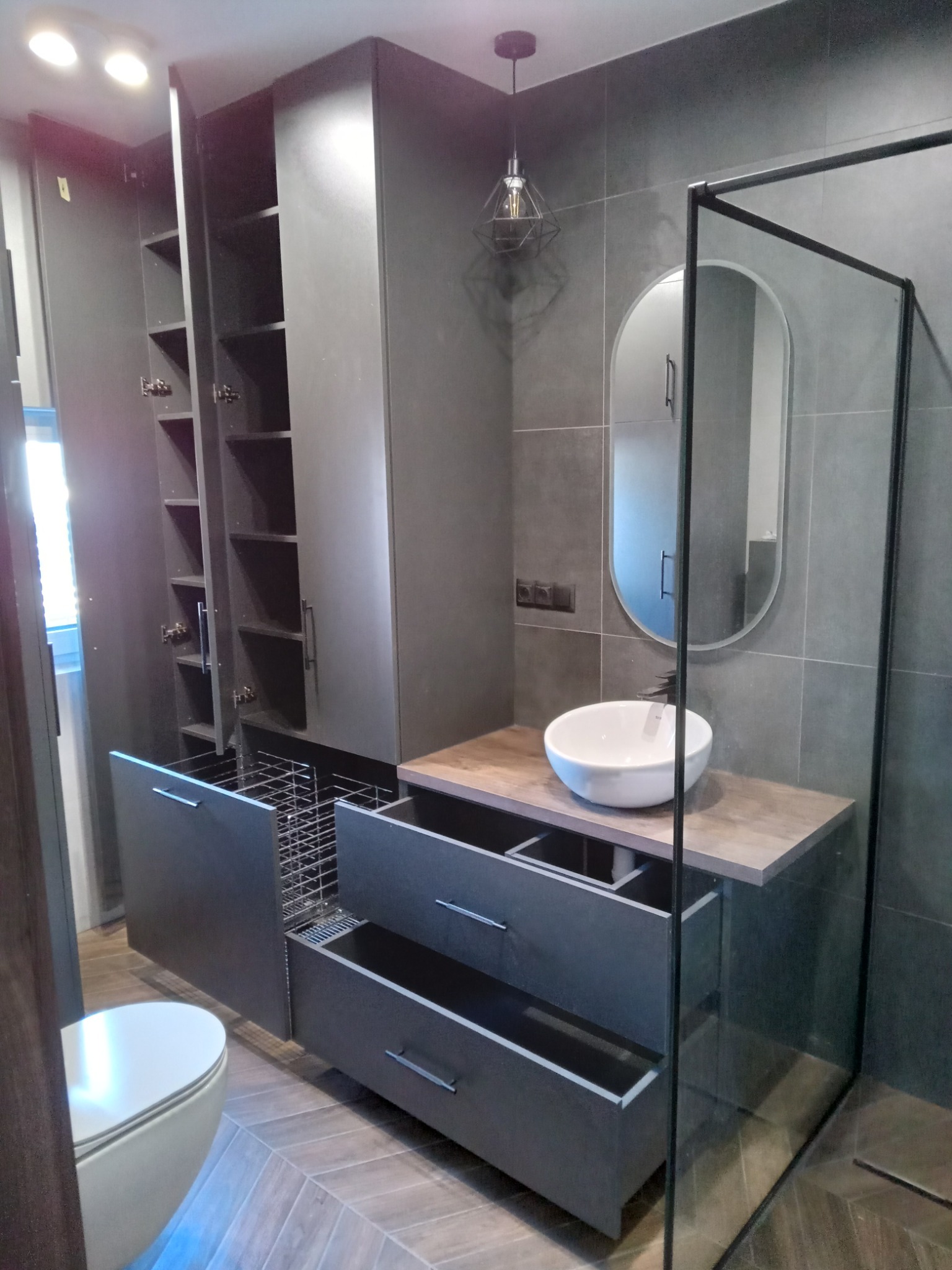 Szara łazienka z szafkami na wymiar, wysuwanymi koszami, umywalką nablatową i kabiną prysznicową typu walk-in, oświetlona lampą wiszącą z geometrycznym kloszem.