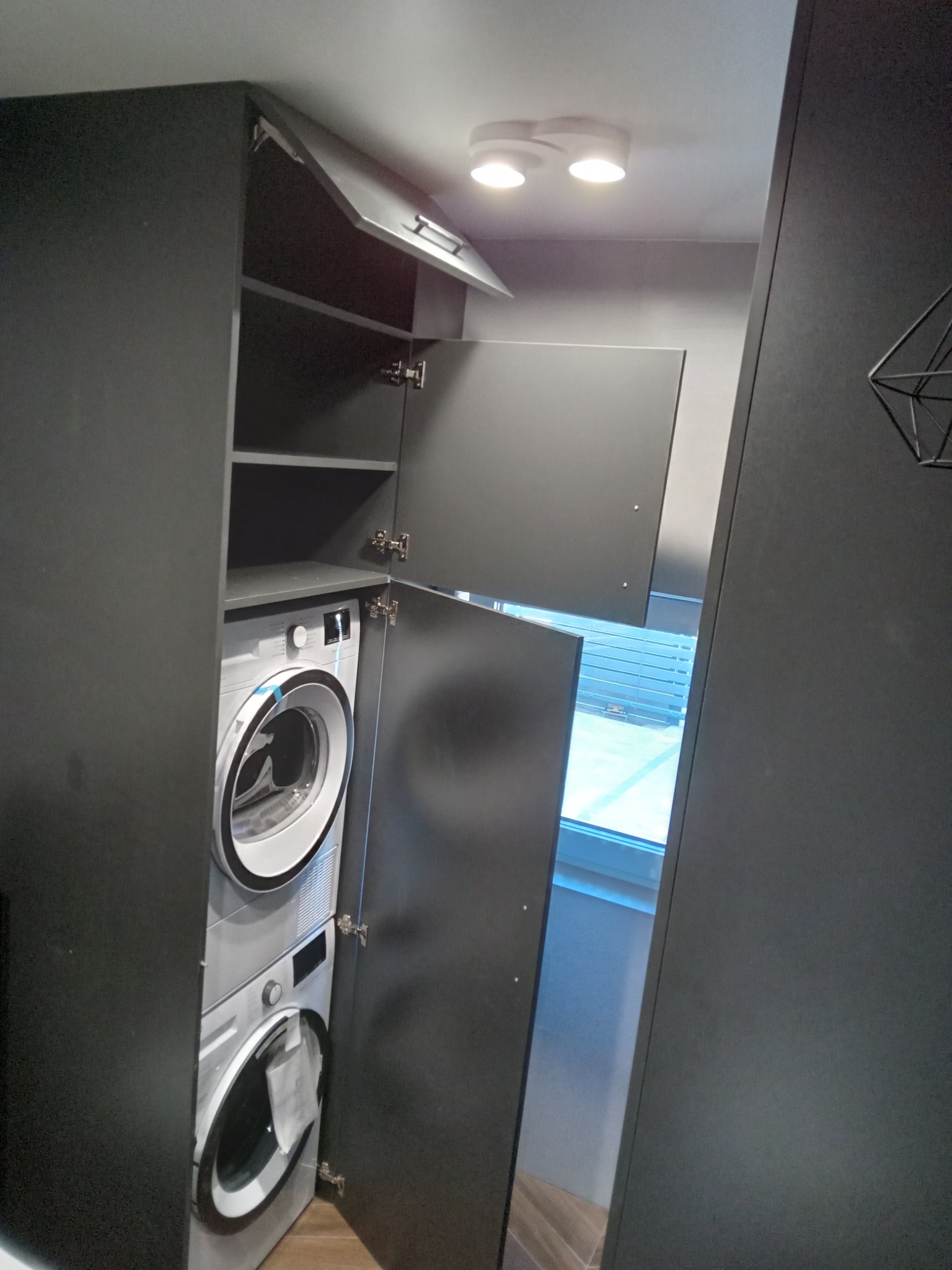 Szara zabudowa meblowa z pralką i suszarką w łazience, drzwi szafki otwarte, widoczne półki i okno z żaluzjami w tle.