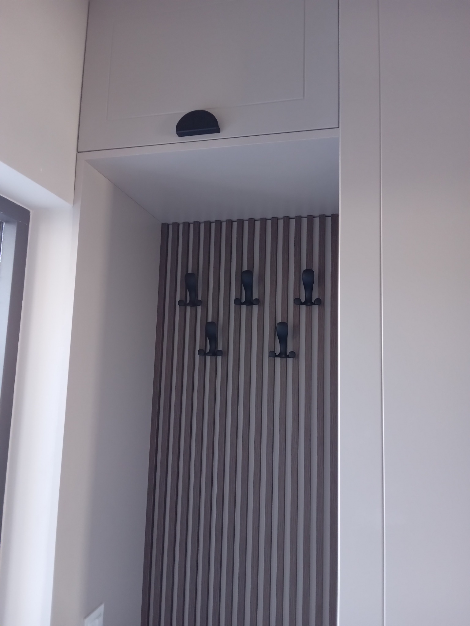 Wnęka z zabudową meblową i panelem lamelowym z wieszakami na ubrania, widok z dołu, jasna kolorystyka.