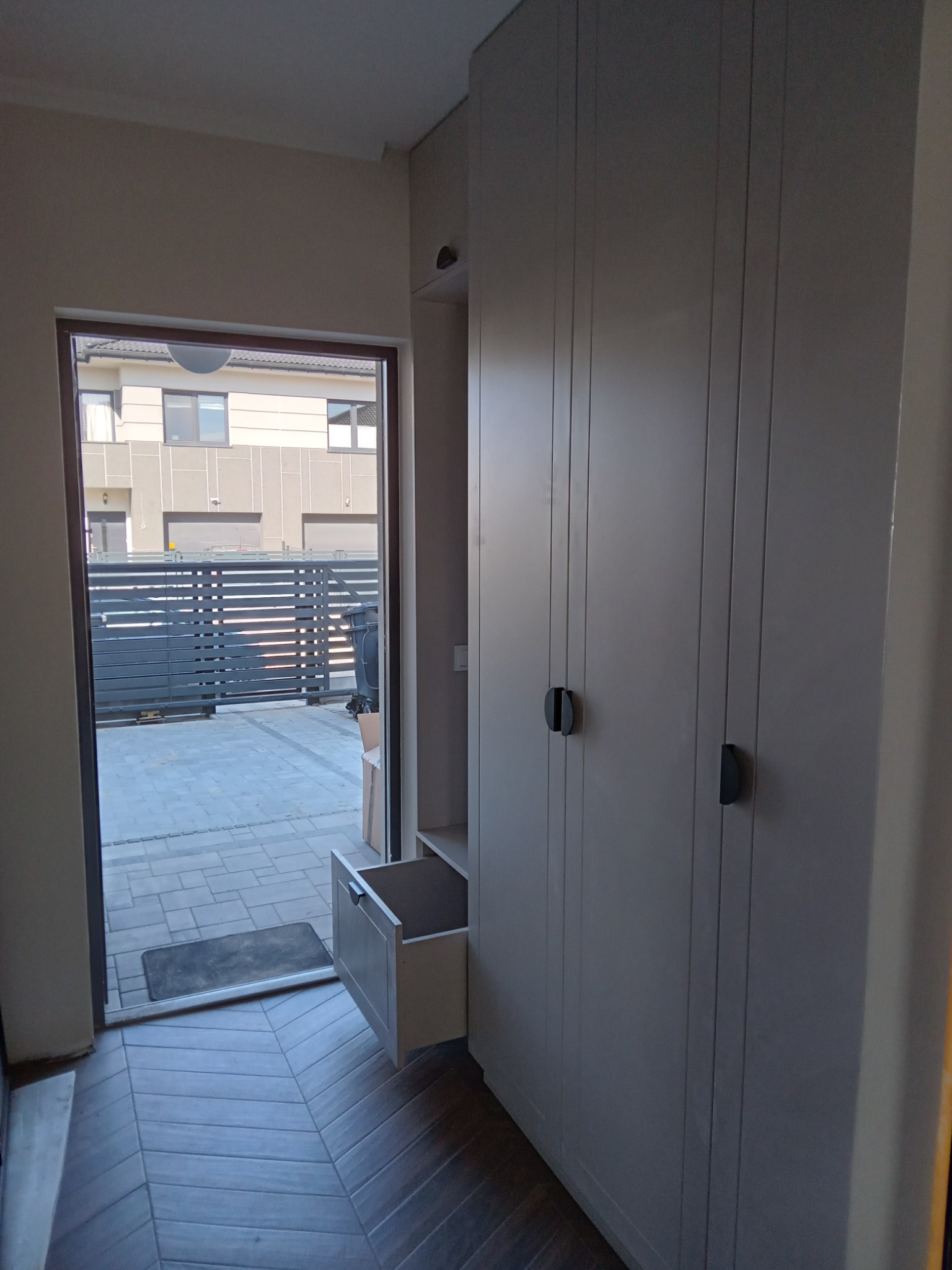 Wysoka, jasnoszara szafa z pionowymi frezowaniami i otwartą szufladą w przedpokoju, widok na zewnątrz przez otwarte drzwi.