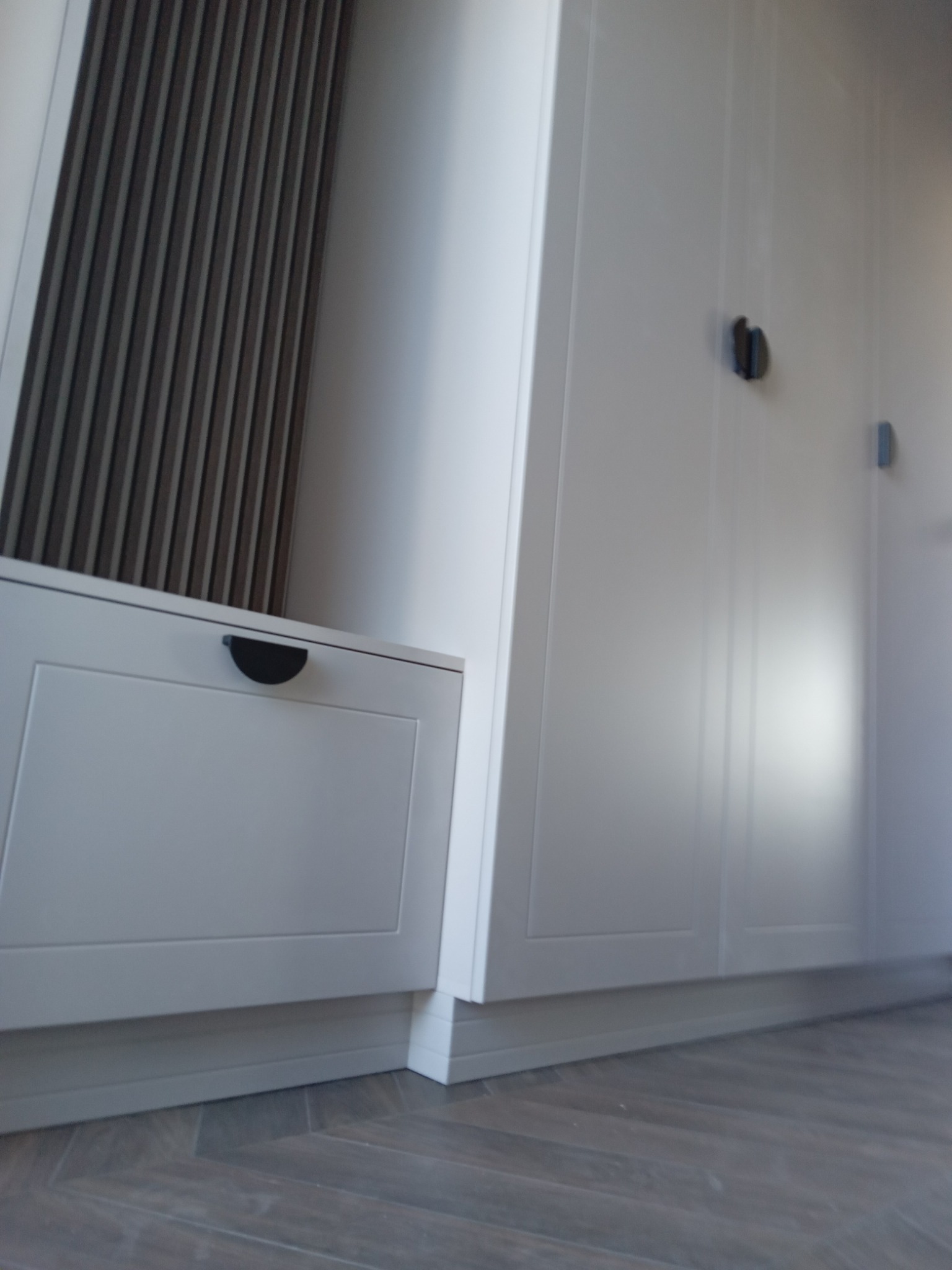 Biała zabudowa meblowa z szufladą i szafami, widoczna z niskiego kąta, z parkietem ułożonym w jodełkę na podłodze i dekoracyjnym panelem ściennym.