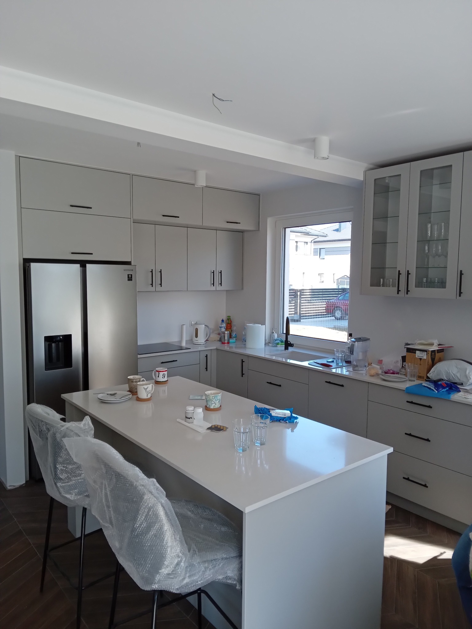 Jasnoszara kuchnia z wyspą, krzesła barowe zabezpieczone folią, widoczne szklanki i naczynia na blacie, nowoczesne wykończenie.