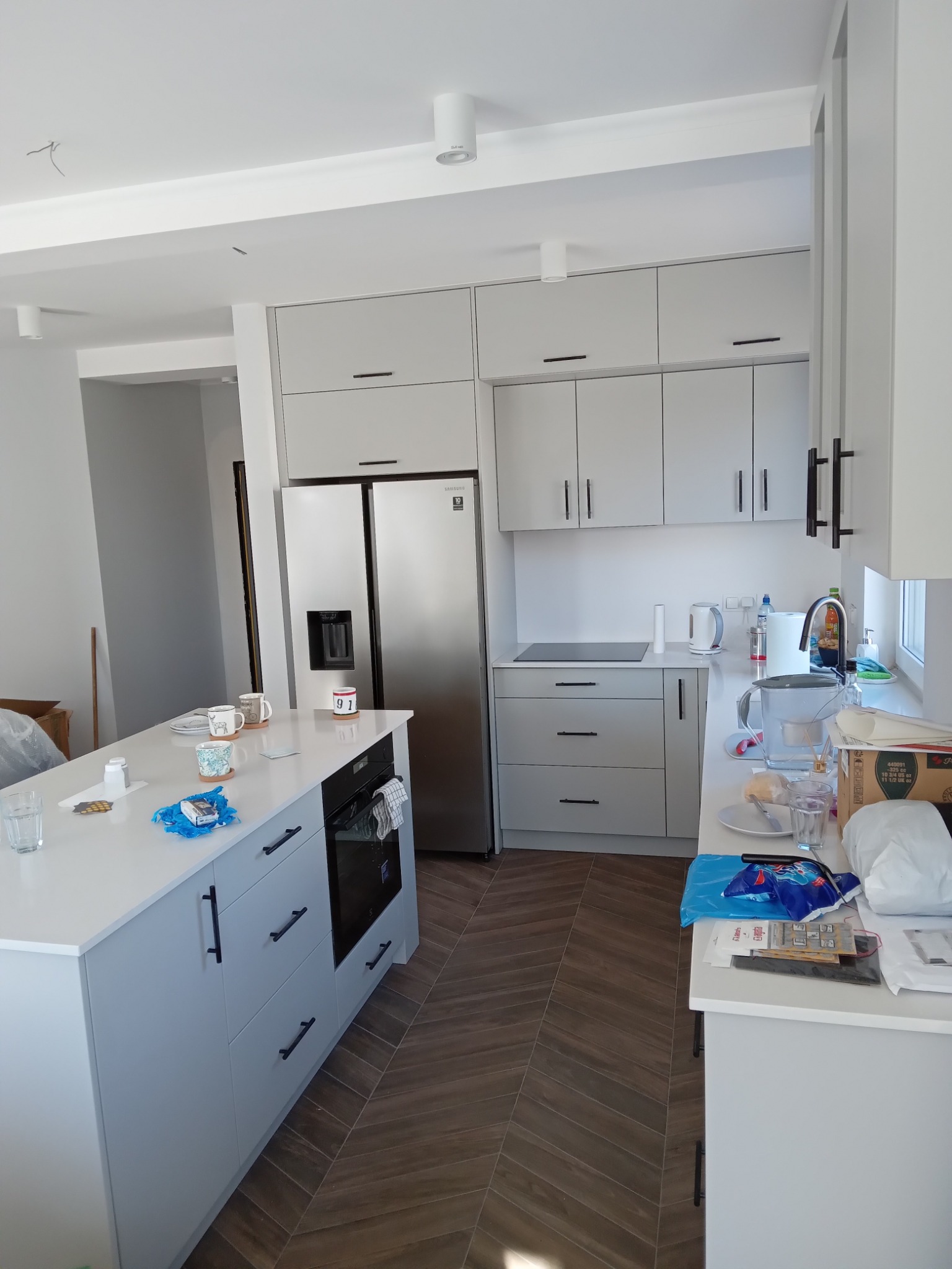 Jasnoszara kuchnia z wyspą centralną i sprzętem AGD, widoczne ślady użytkowania i niedawnego montażu, na blacie kubki, naczynia i akcesoria, podłoga wyłożona parkietem w jodełkę.