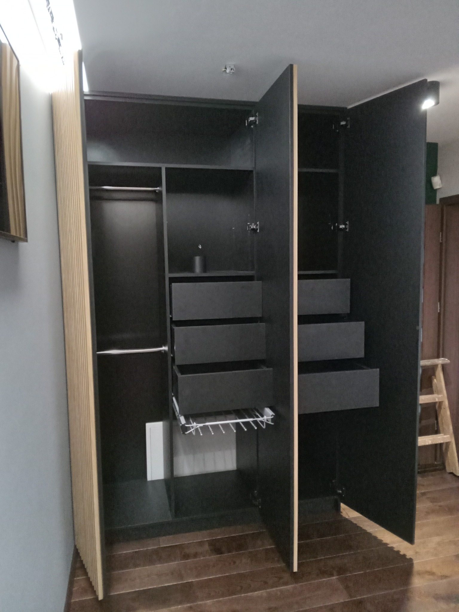 Czarna szafa z szufladami i drążkami na ubrania, z jasnymi, pionowo żłobionymi bokami, zrealizowana w Stalowej Woli. Częściowo otwarte drzwi ukazują wnętrze.