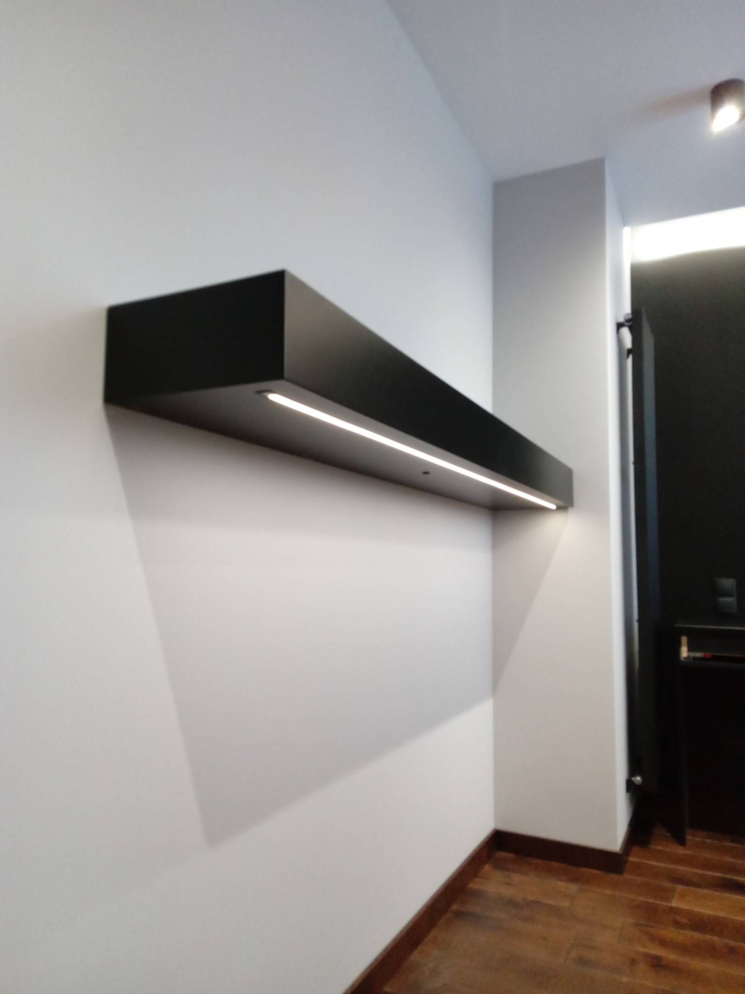 Czarna, podświetlana półka zamontowana na białej ścianie w nowoczesnym wnętrzu, widok z dołu, podłoga drewniana
