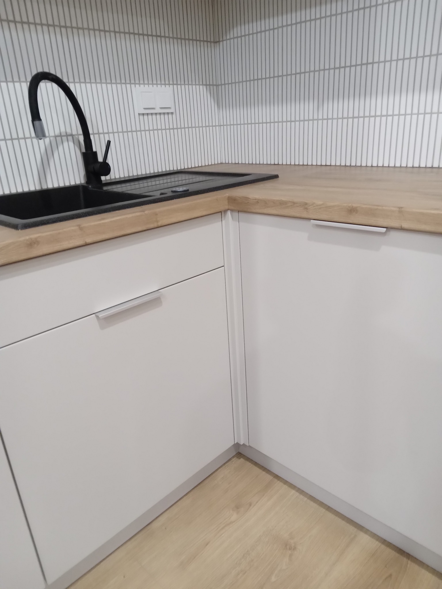 Nowoczesna kuchnia z białymi szafkami, drewnianym blatem i czarnym zlewem z baterią, minimalistyczne płytki na ścianie.