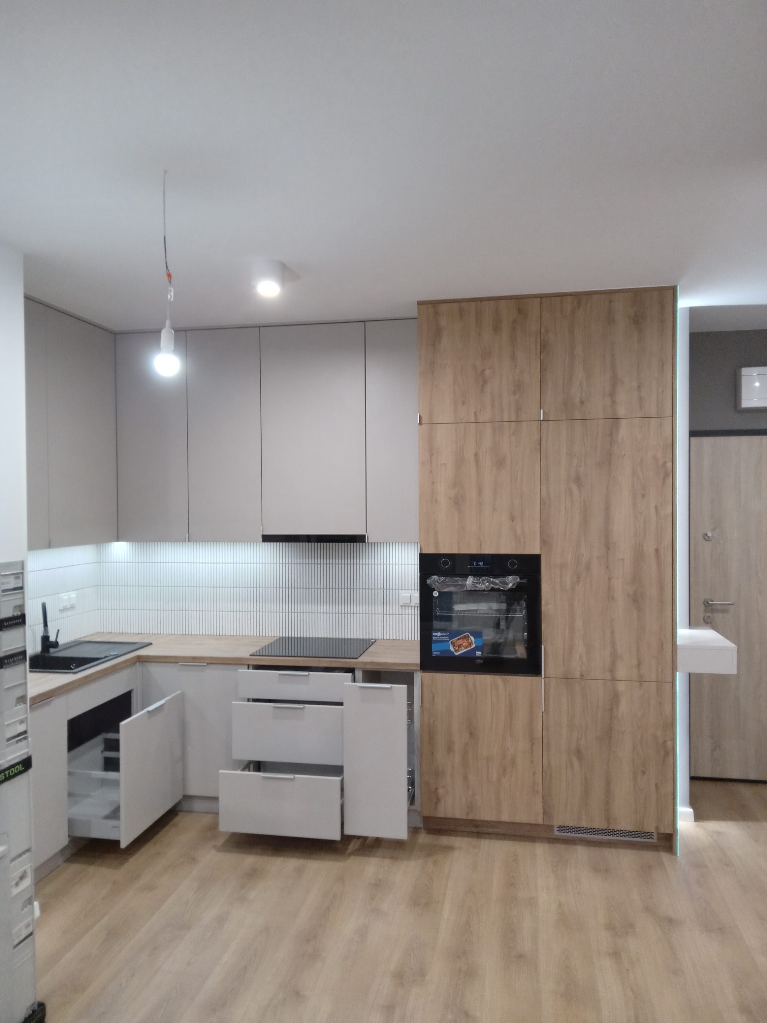 Nowoczesna kuchnia z frontami w odcieniach szarości i drewna, otwarte szuflady eksponują wewnętrzne przegródki, a białe płytki nad blatem dodają subtelności.