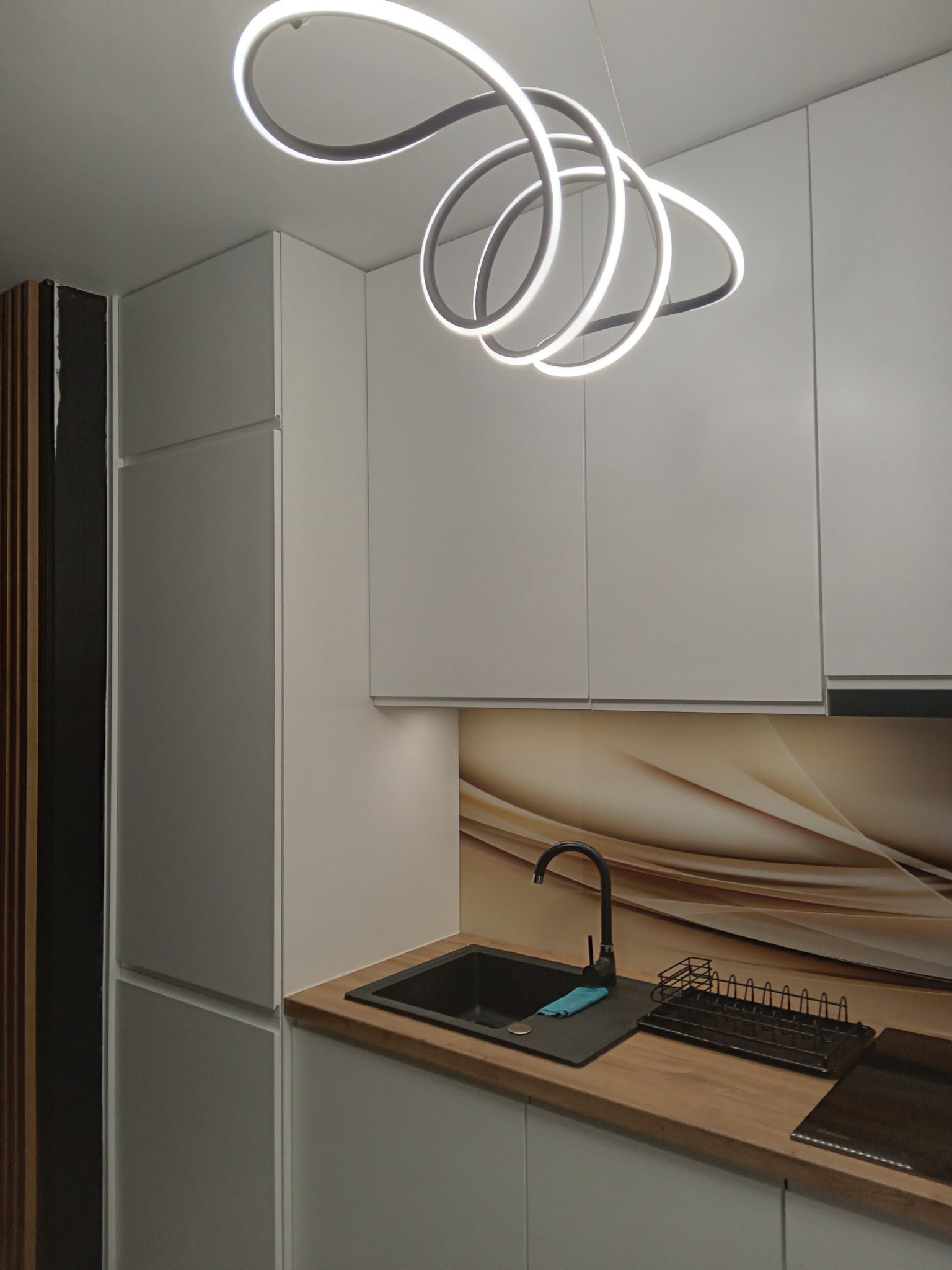 Nowoczesna kuchnia z białymi szafkami, czarnym zlewem i baterią, drewnianym blatem oraz designerską lampą sufitową w kształcie spirali.