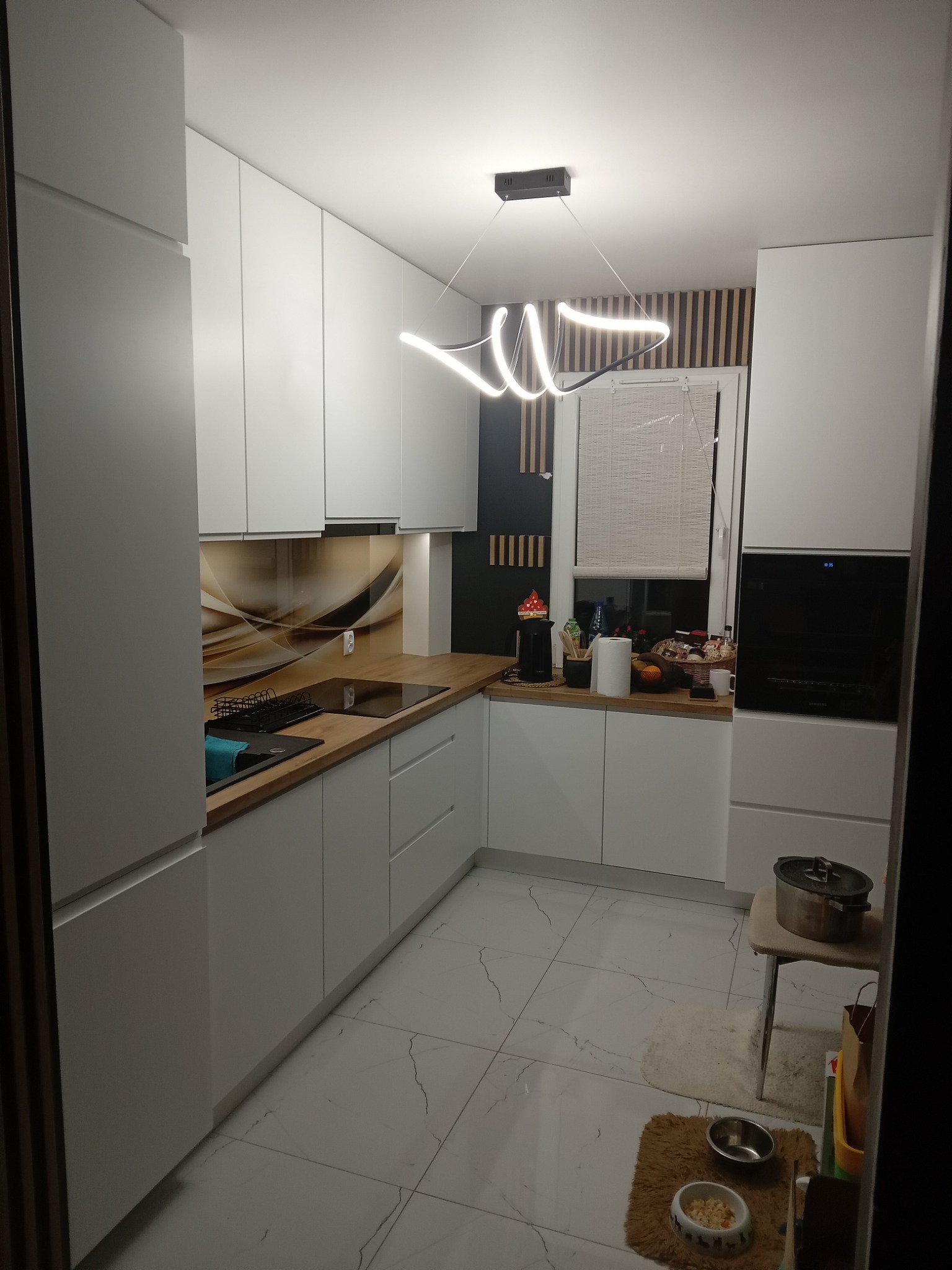 Nowoczesna kuchnia z białymi szafkami, drewnianymi blatami i abstrakcyjną lampą LED, widok na okno z żaluzjami.