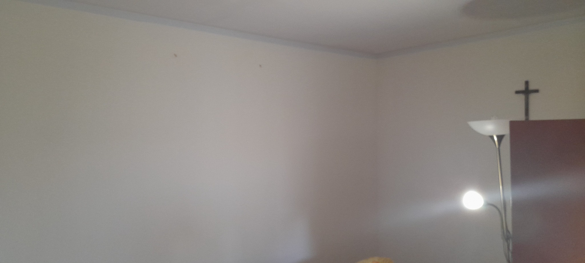 Pomalowana na jasny kolor ściana w rogu pokoju z widocznymi śladami po wkrętach, listwa przysufitowa w odcieniu szarości, lampa podłogowa i krzyż na szafce w tle.
