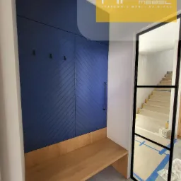 Nowoczesna garderoba z niebieskimi frontami szafy zdobionymi ukośnymi liniami, drewnianym siedziskiem i czarnymi haczykami, widok na klatkę schodową przez szklaną ścianę z czarną ramą.