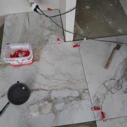 Dagmara Tarasewicz - Układanie marmurowych płytek podłogowych z użyciem systemu poziomowania, widoczne narzędzia: młotek, przyssawka, krzyżyki dystansowe, poziomica, okulary i wiadro z klipsami.