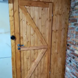 Rustykalne drewniane drzwi z ukośnym wzmocnieniem, czarną gałką i metalową klamką, sąsiadujące z białą ścianą i fragmentem ściany z czerwonej cegły.