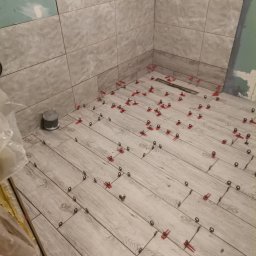 Dagmara Tarasewicz - Układanie płytek imitujących drewno na podłodze w łazience w Szemudzie, z użyciem czerwonych klipsów poziomujących. Częściowo wykończone ściany z szarych, kwadratowych płytek.