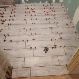 Dagmara Tarasewicz - Układanie płytek podłogowych imitujących drewno z użyciem klipsów poziomujących, widoczne niedokończone ściany i fragment drewnianej podłogi w tle.