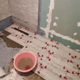 Układanie płytek imitujących drewno w łazience, widoczne czerwone klipsy poziomujące, wiadro z brudną wodą i paczka klipsów, ściany częściowo wykończone, częściowo surowe.