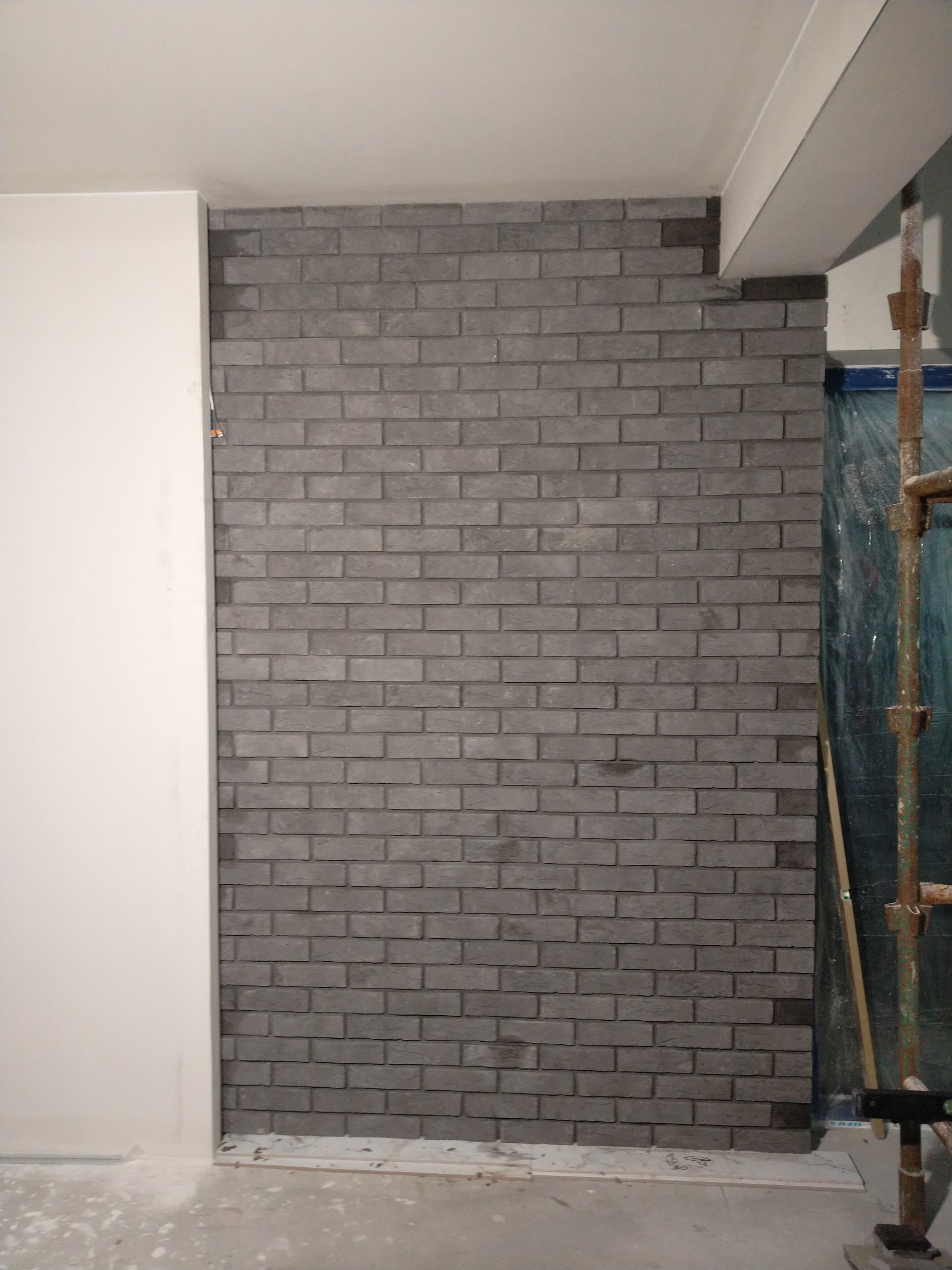 Ściana wykończona szarą cegłą dekoracyjną, widoczne fragmenty rusztowania i zabezpieczona folią przestrzeń, surowy stan wykończenia podłogi.