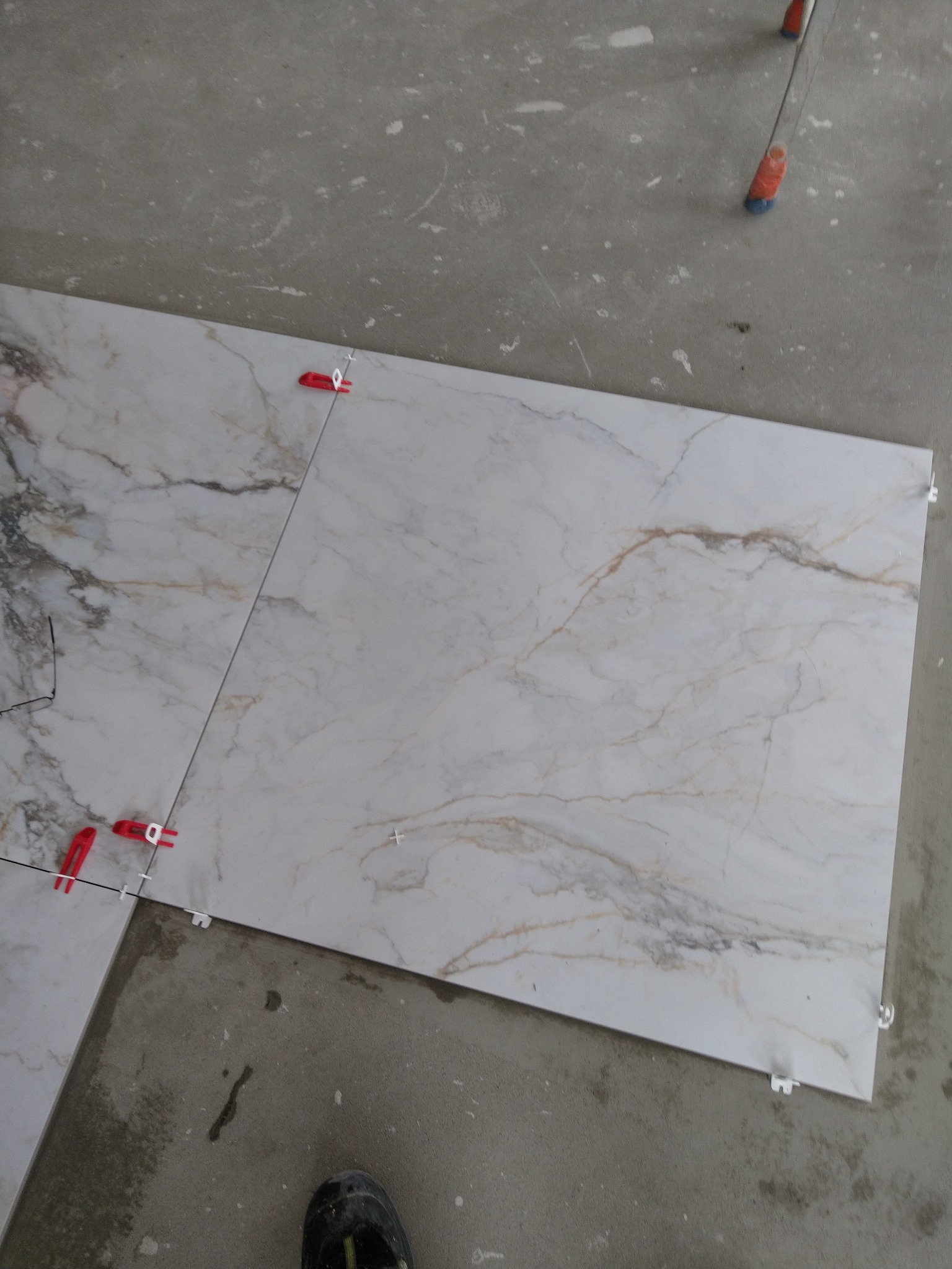 Układanie płytek imitujących marmur na podłodze, z widocznymi czerwonymi i białymi klipsami poziomującymi, na surowym, szarym podłożu.